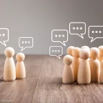 Holzfiguren mit Sprechblasen symbolisieren Austausch zwischen Gruppen und veranschaulichen die Bedeutung von Kommunikation im Berufsalltag.